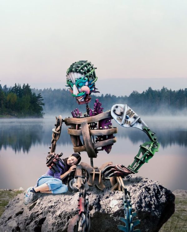 A giant puppet sitting on a rock on the shore of a lake, with a performer resting on its side.
