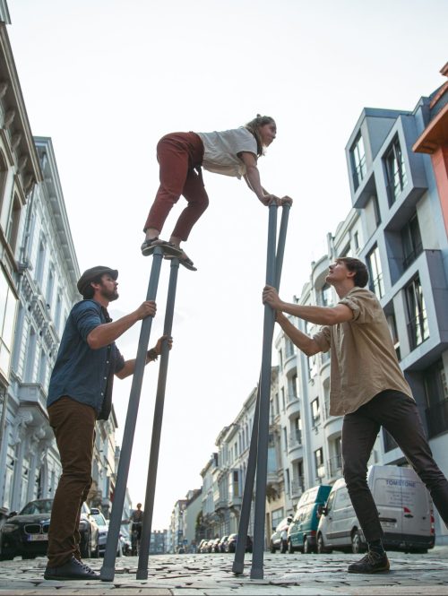 Three performers on a street, with one of them balancing in the air on sticks.