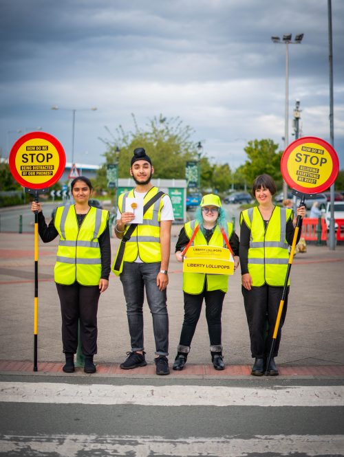 Artist Davina and three Changemakers stand at a crossroads, wearing yellow high-vis and lollipop signs