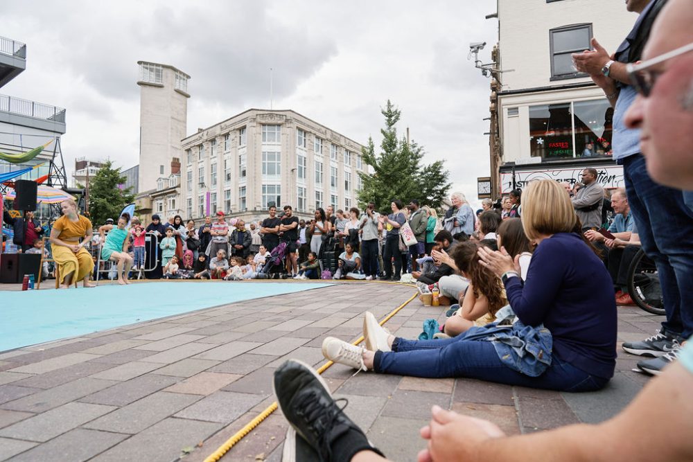 Audiences sitting on the floor and standing around an outdoor stage around Leicester's Clock Tower.