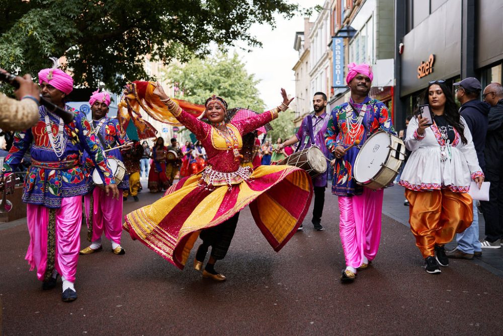 A woman in colourful and traditional Indian costume. The woman is dancing to live music by drummers who are also in the image.