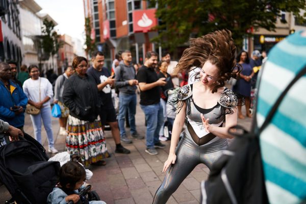 A performer in costume dancing on an outdoor square, surrounded by audiences.