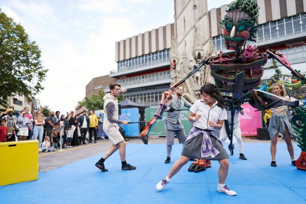 A young woman person is dancing at an outdoor arts festival with a giant puppet behind her and members of the public in the background at Leicester's Clock Tower.