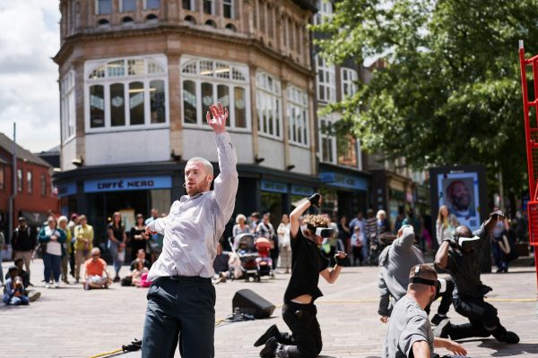 A group of dancers, some wearing VR headsets, are performing in front of a crowd on a high street at an outdoor arts festival.