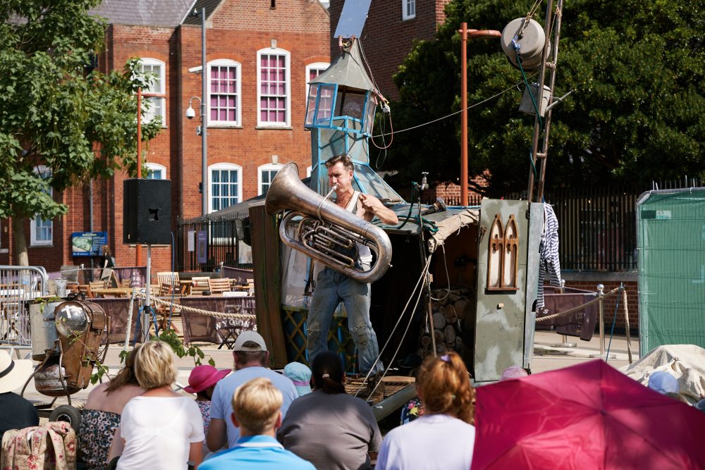 This image shows a man performing in front of an audience. The set looks like small caravan on a farm and the man is playing a huge brass instrument.