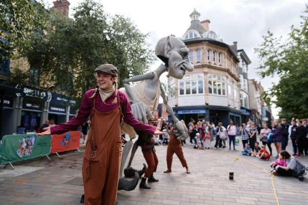A giant grey puppet is on a busy high street and an actor stands in the foreground.