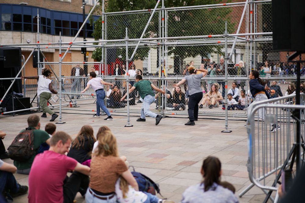 Performers dancing next to a scaffolding structure on an outdoor square, with audiences around watching them.