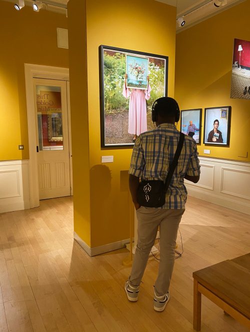 A young man is wearing earphones and looking at a picture in a gallery. The picture is hung on a yellow wall and looks like a painting of a person holding a pinting in front of their face.