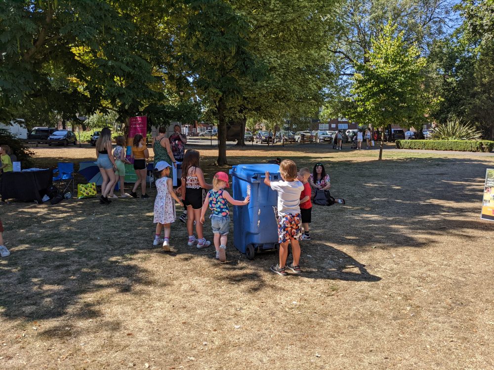 A group of children looking at a blue wheelie bin in a park.