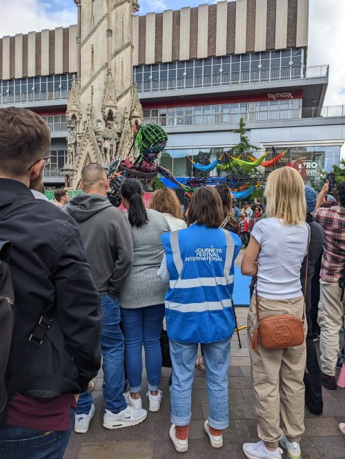 A team member in a blue-vis vest standing amongst other audience members on an outdoor square.