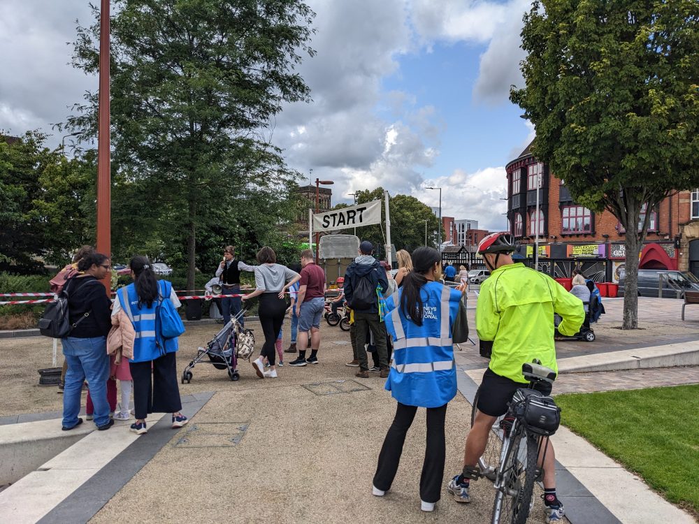 Two festival team members in blue-vis vests talking to audience members on an outdoor square.