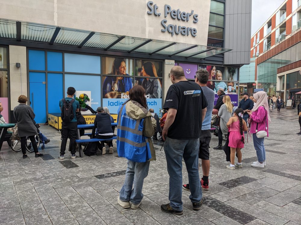 A team member in a blue-vis vest talking to an audience member on an outdoor square.