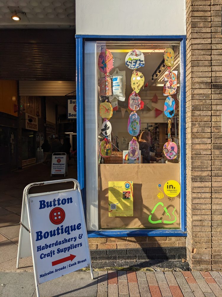Giant sequins displayed hanging in a window at the Button Boutique, with some colourful illustrations of the recycling sign on the bottom, and a sign saying Button Boutique next to the window.