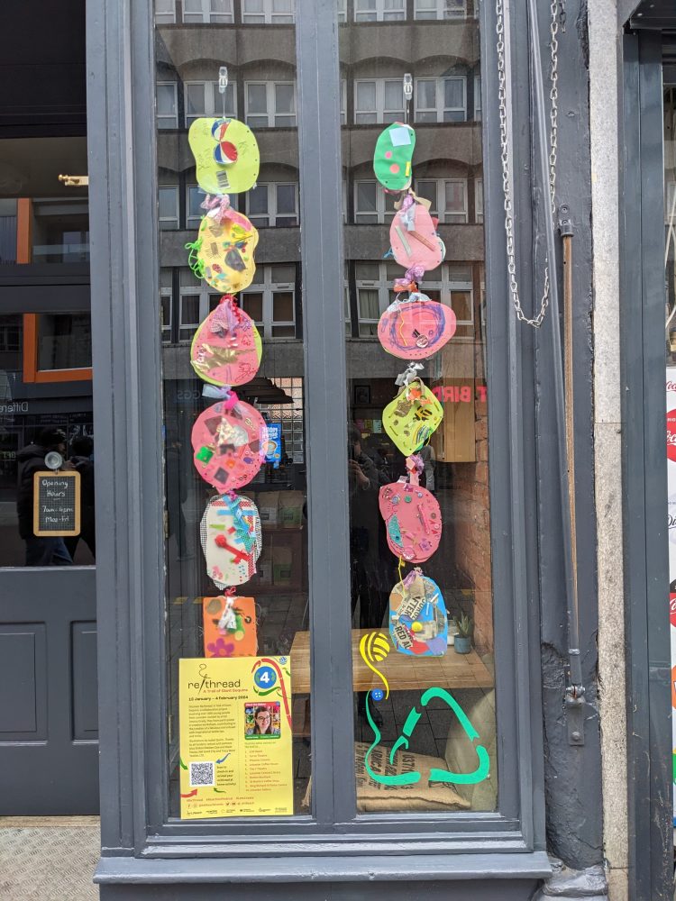 Giant sequins displayed hanging in a window at Leicester Coffee House, with some colourful illustrations of the recycling sign on the bottom.