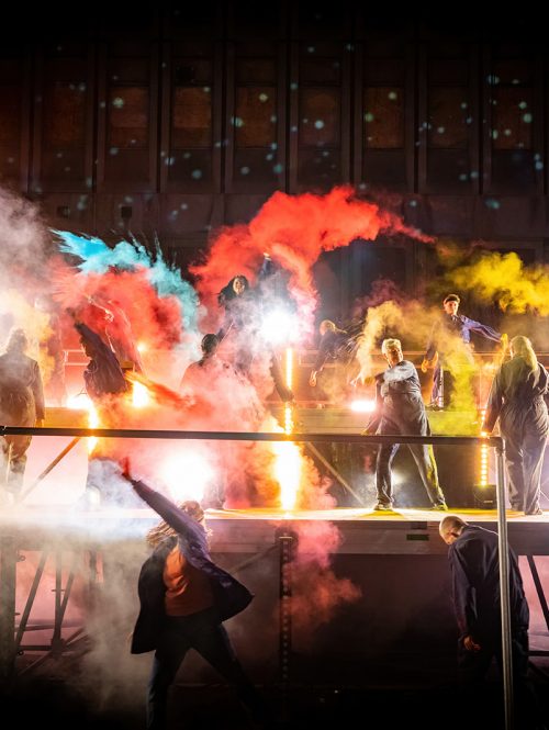 Performers on an outdoor stage in the evening throwing coloured dust into the air.