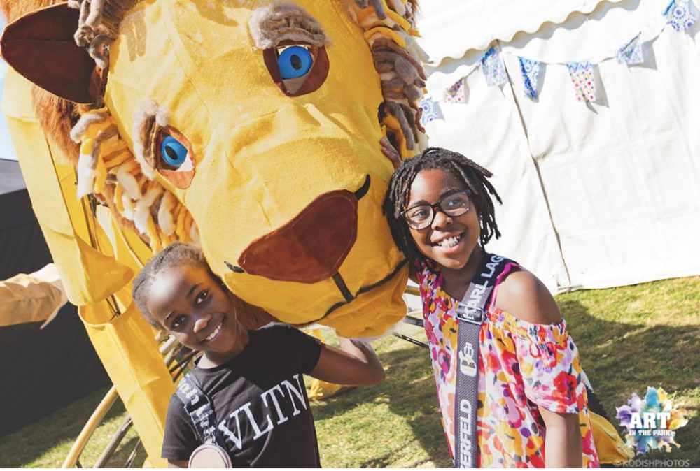 Two young children are in front of a white tent with a huge carnival puppet in the shape of a lion's head in between them.