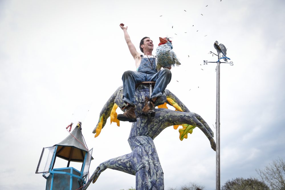 A performer sits on top of an artificial tree with the puppet of a bird in his hand.