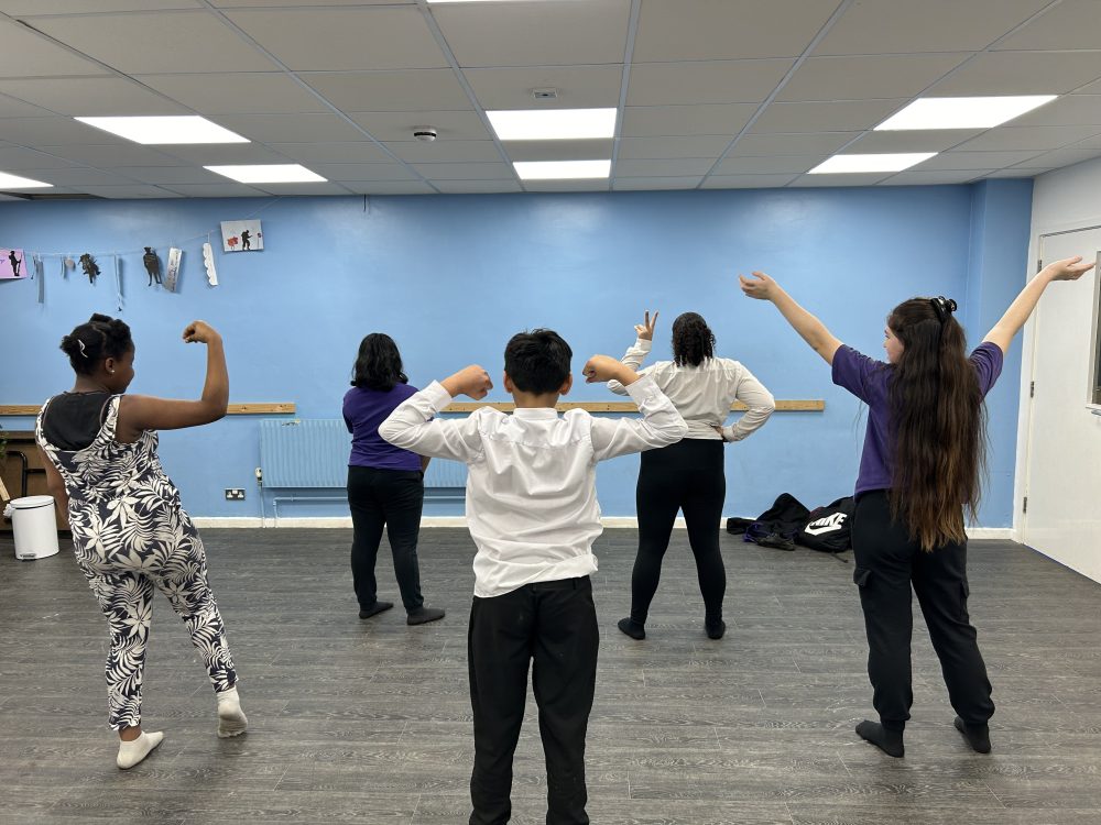 A group of young people are in a rehearsal space which has a big blue wall. They are all facing away from camera and striking a power pose.