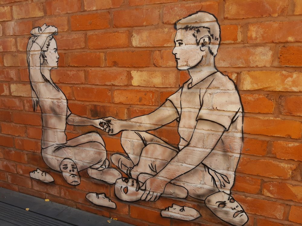 This image shows an outdoor painting by The Rebel Bear at The Queen of Bradgate in Leicester. On the red brick wall you can see a black and white painting of a couple sitting down, holding hands. There are also some comedy and tragedy theatre masks scattered around the couple.