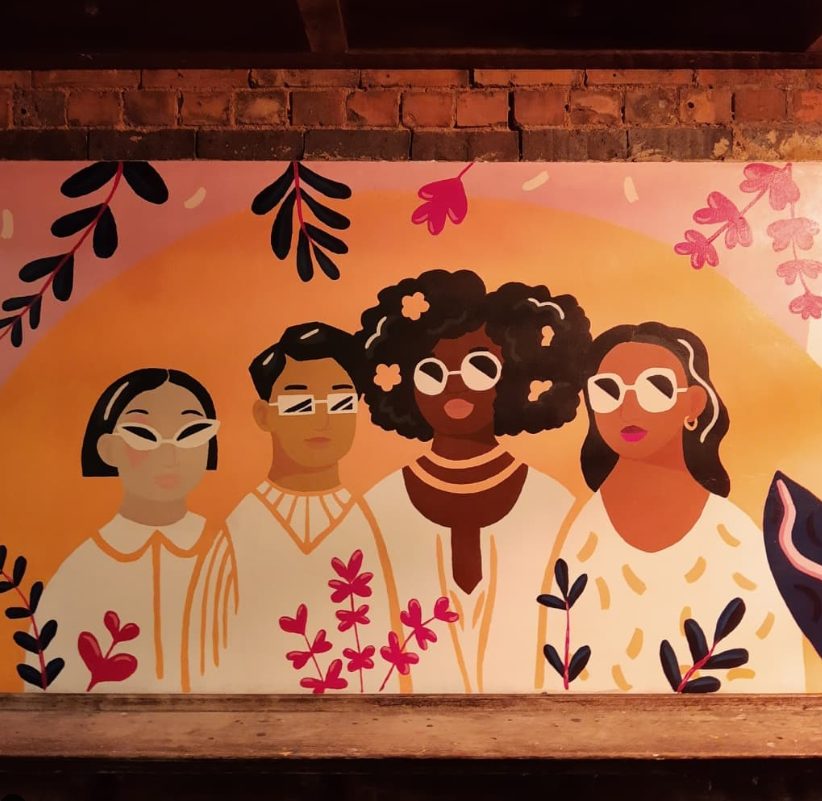 A painting of four people with different skin colours wearing sunglasses and white clothes surrounded by flowers and plants on an exterior brick wall.