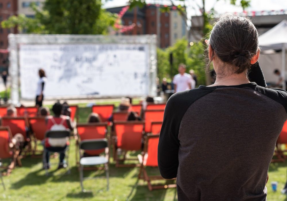 The back of a person looking at an outdoor stage with red canvas chairs for audiences.