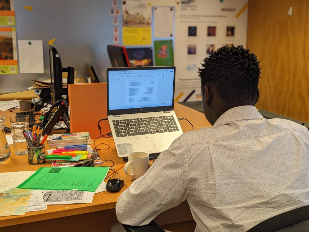 A young person sitting at a desk in an office space, writing on a laptop.