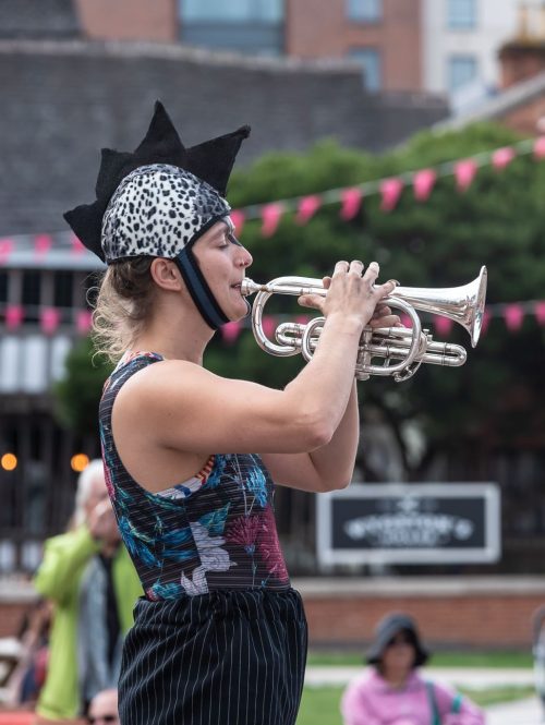 A woman stands on stilts in a wild costume and plays the trumpet in front of an audience.