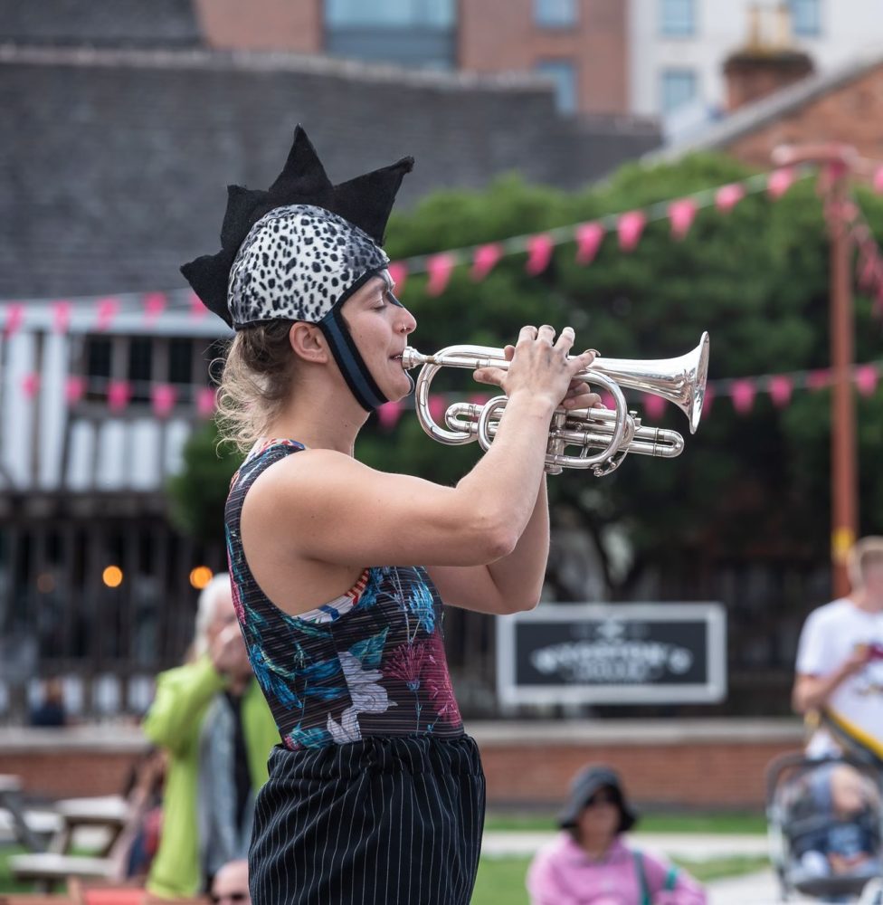 A woman stands on stilts in a wild costume and plays the trumpet in front of an audience.