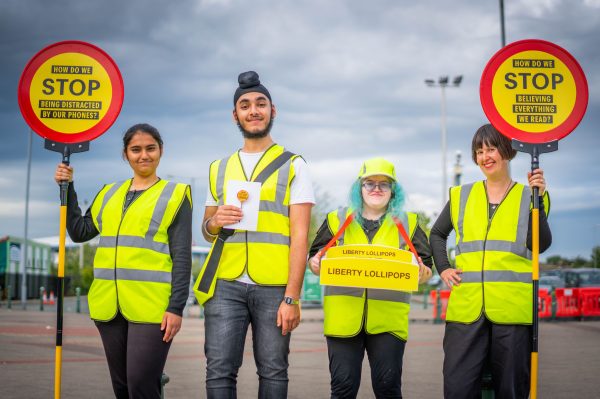 This image shows four people of varying age and ethnicity that are all wearing, what appears to be, a school crossing wardens uniform. They are holding trays of lollipop sweets and two of them are holding lollipop signs. One sign reads, How do we stop being distracted by our phones, the other reads, How do we stop believing everything we read.