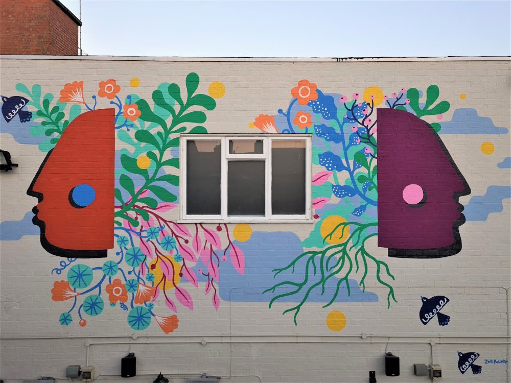 This is an image of an outdoor painting by Zoe Power at The Rutland and Derby in Leicester. The painting is on a big wall with a window in the middle. The background is all white and features colourful flowers, plants and birds. The main focus of the painting is two faces in profile, one is purple and one is orange, the have no facial features but do have a contrasting coloured dot on them.